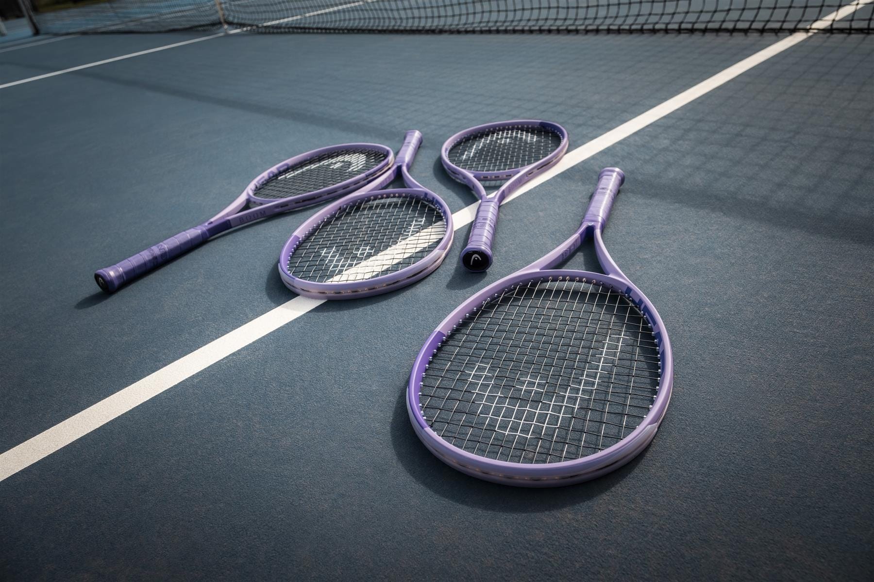 Four purple HEAD Boom MP L Alternate 2026 tennis rackets from HEAD rest on a blue court near the white boundary line, with the net in view.