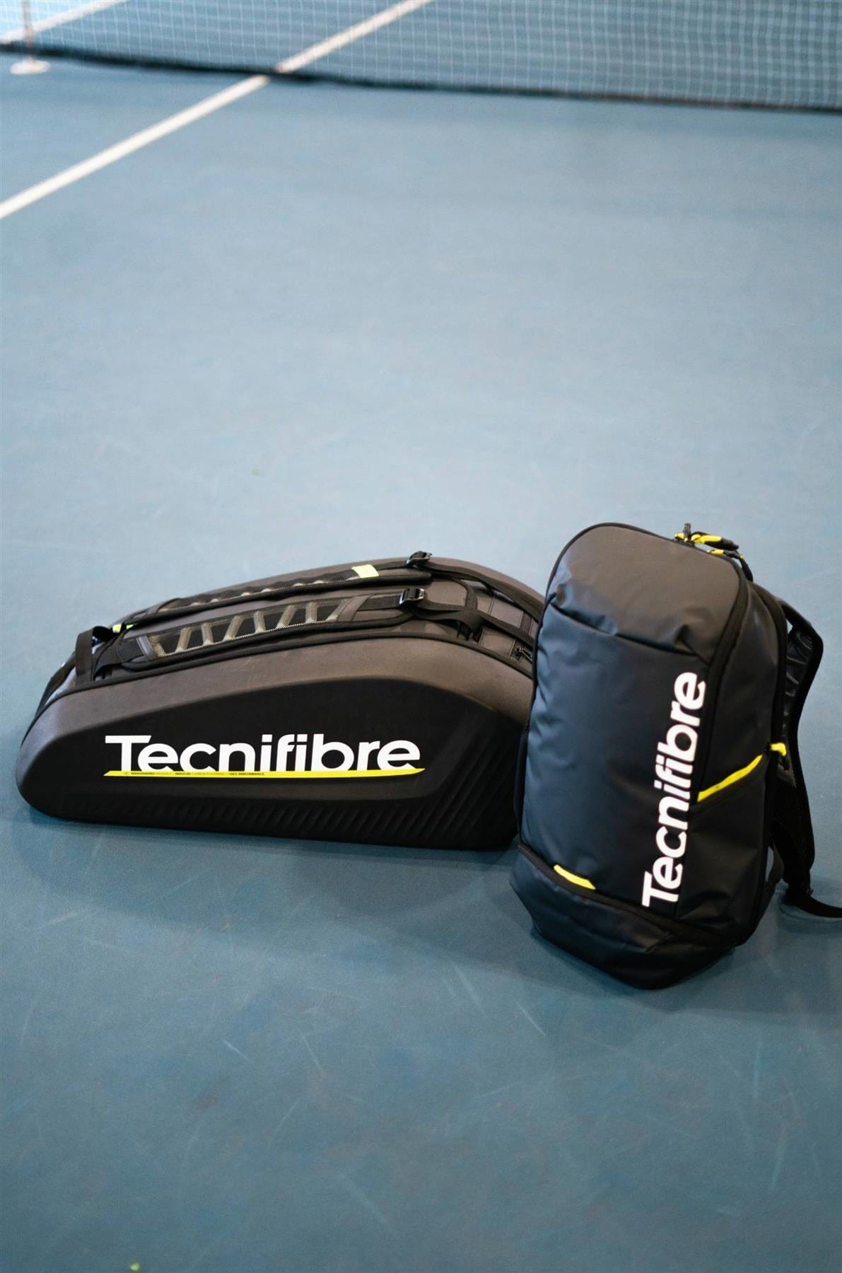 Two black Tecnifibre Reform L Tennis Racket Bags rest on a blue tennis court near the net. Designed for durability, each bag features a white Tecnifibre logo, yellow accents, and spacious compartments for tennis gear.