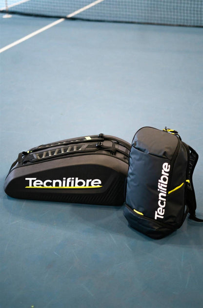 Two black Tecnifibre Reform L Tennis Racket Bags rest on a blue tennis court near the net. Designed for durability, each bag features a white Tecnifibre logo, yellow accents, and spacious compartments for tennis gear.
