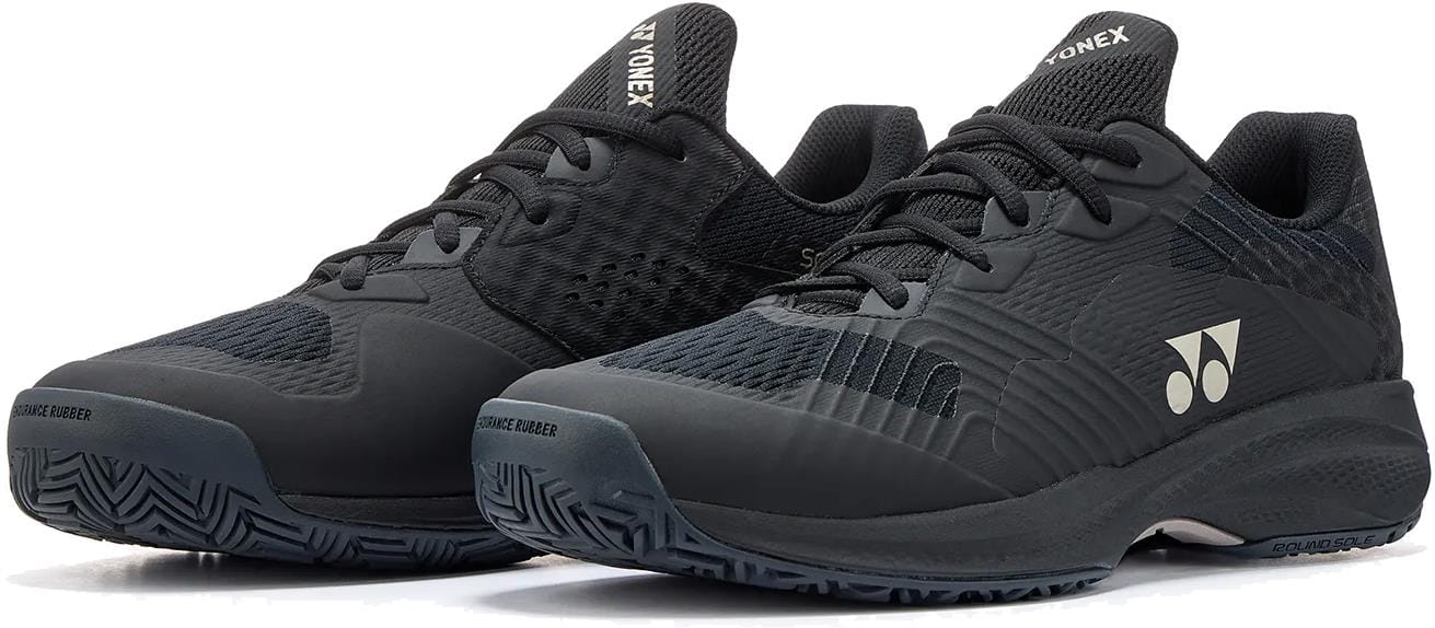 A pair of Yonex Power Cushion Sonicage Men's Tennis Shoes in Black/Mocha Grey, featuring textured rubber soles, mesh panels for breathability, and Yonex logos on the sides and tongue, shown side by side on a white background. Durable and lightweight.