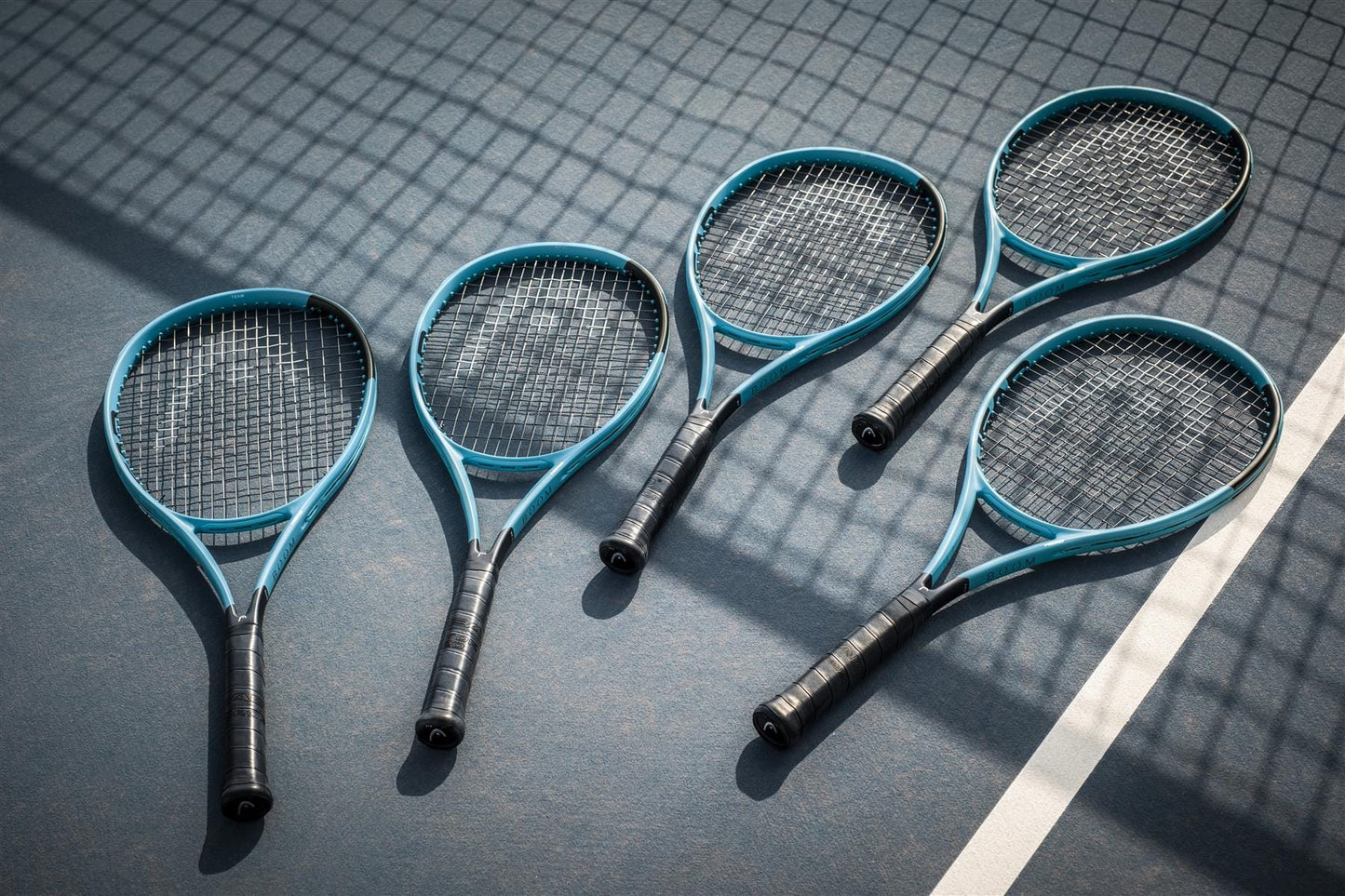 Five HEAD Boom MP L 2026 tennis rackets, featuring a sleek blue and black design, are arranged side by side on the court near a white line, with shadows from the net highlighting their power and performance.