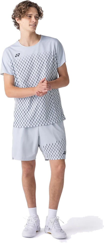A young man wears the Yonex 10713EX Crew Neck Men's Tennis T-Shirt in Silver Grey, paired with shorts featuring a dotted black pattern and white sneakers. He stands looking to the side, clasping his hands together.