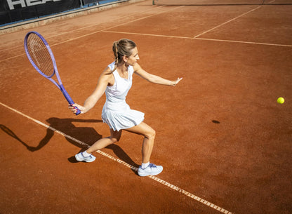 On a sunny clay court, a woman in white gets ready to smash a yellow ball with her HEAD Boom MP Alternate 2026 Tennis Racket, channeling explosive power into every shot.