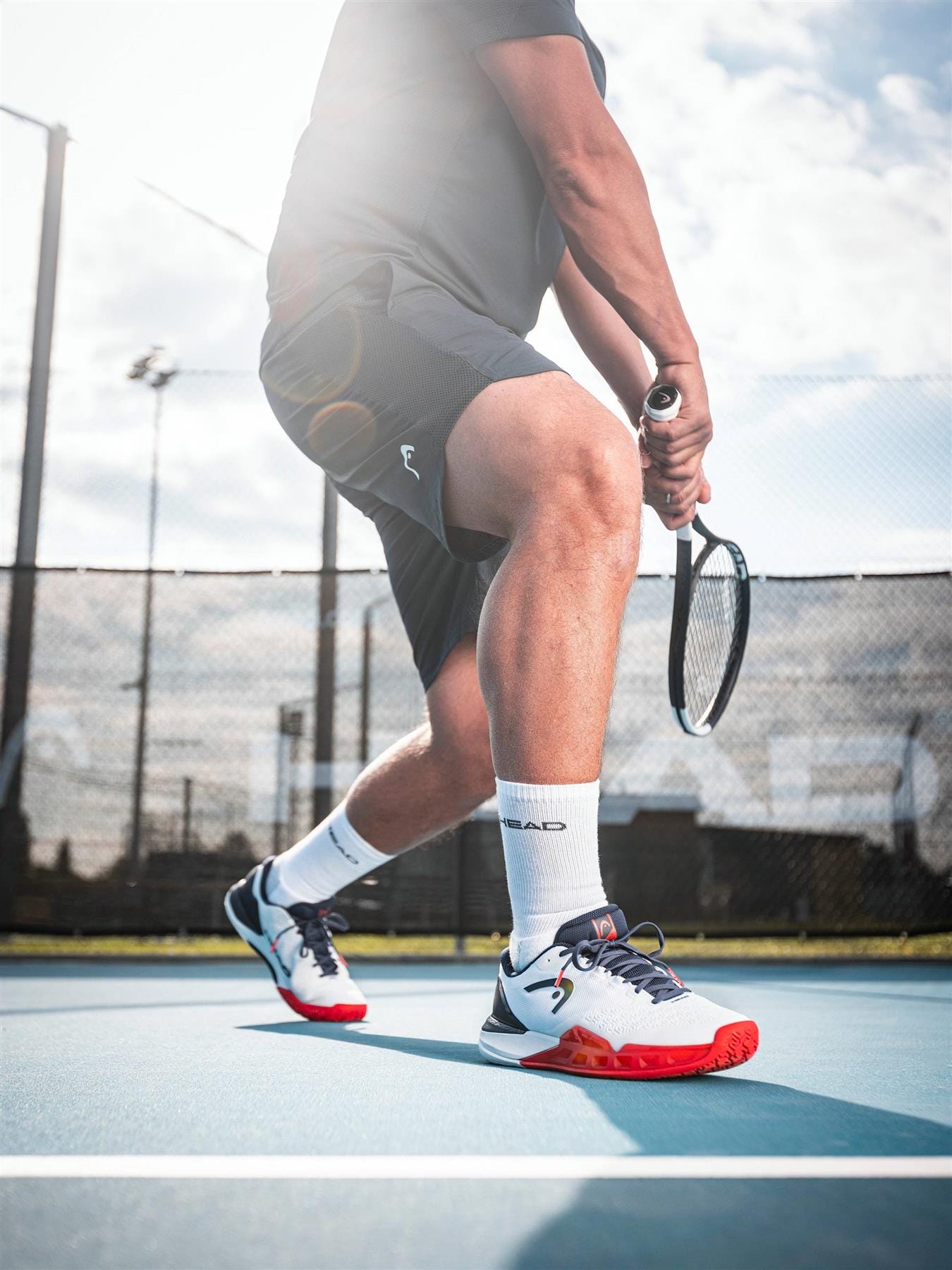A tennis player in black shorts and HEAD Revolt Pro 5.0 Mens Tennis Shoes in White/Navy readies a backhand on an outdoor court, sunlight streaming behind him and a fence visible—an iconic moment for baseline players.