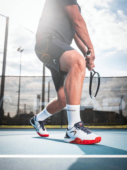 A tennis player in black shorts and HEAD Revolt Pro 5.0 Mens Tennis Shoes in White/Navy readies a backhand on an outdoor court, sunlight streaming behind him and a fence visible—an iconic moment for baseline players.