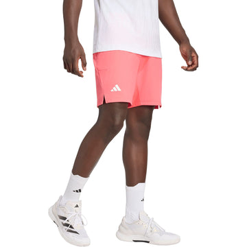 A person wearing a white shirt, adidas Ergo 9 Inch Mens Tennis Shorts - Pure Turbo featuring HEAT.RDY technology, white adidas socks, and white sneakers stands against a plain white background.