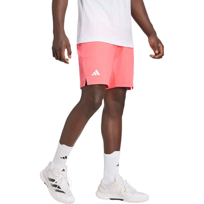 A person wearing a white shirt, adidas Ergo 9 Inch Mens Tennis Shorts - Pure Turbo featuring HEAT.RDY technology, white adidas socks, and white sneakers stands against a plain white background.