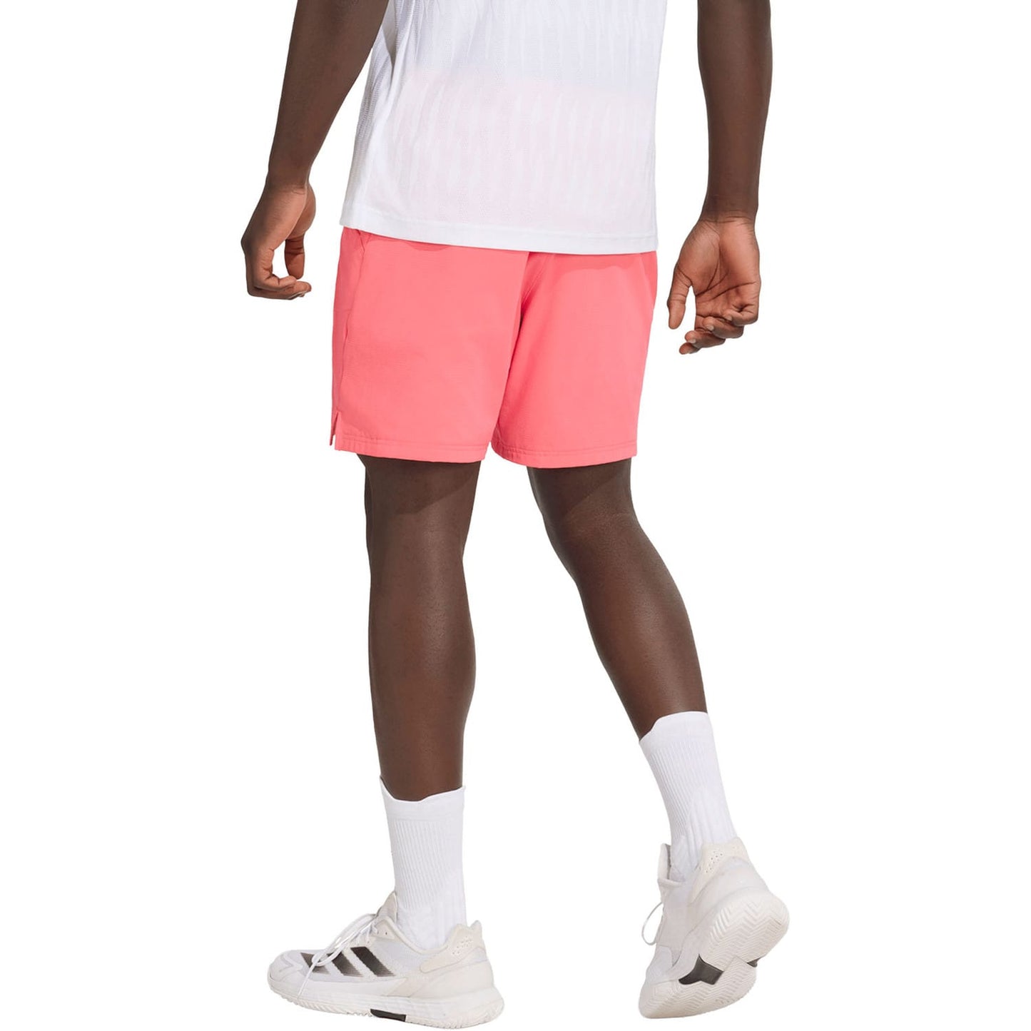 A person wearing a white shirt, bright pink adidas Ergo 9 Inch Mens Tennis Shorts in Pure Turbo, white crew socks, and white sneakers stands facing away from the camera against a white background.
