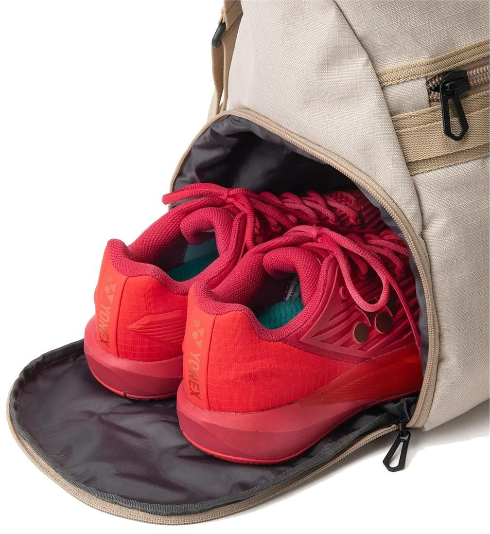 A pair of bright red Yonex athletic shoes are partially tucked into the open zippered shoe compartment of a beige Yonex BA72611EX Gearlogic Tennis Boston Bag, with their soles facing outward.