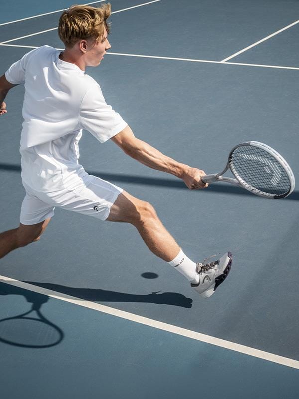 A male tennis player in white lunges for a backhand on a blue outdoor court, gripping his HEAD Squared Tennis Racket from HEAD, which features an ultra-head light design and dual-tube technology as his shadow trails behind.