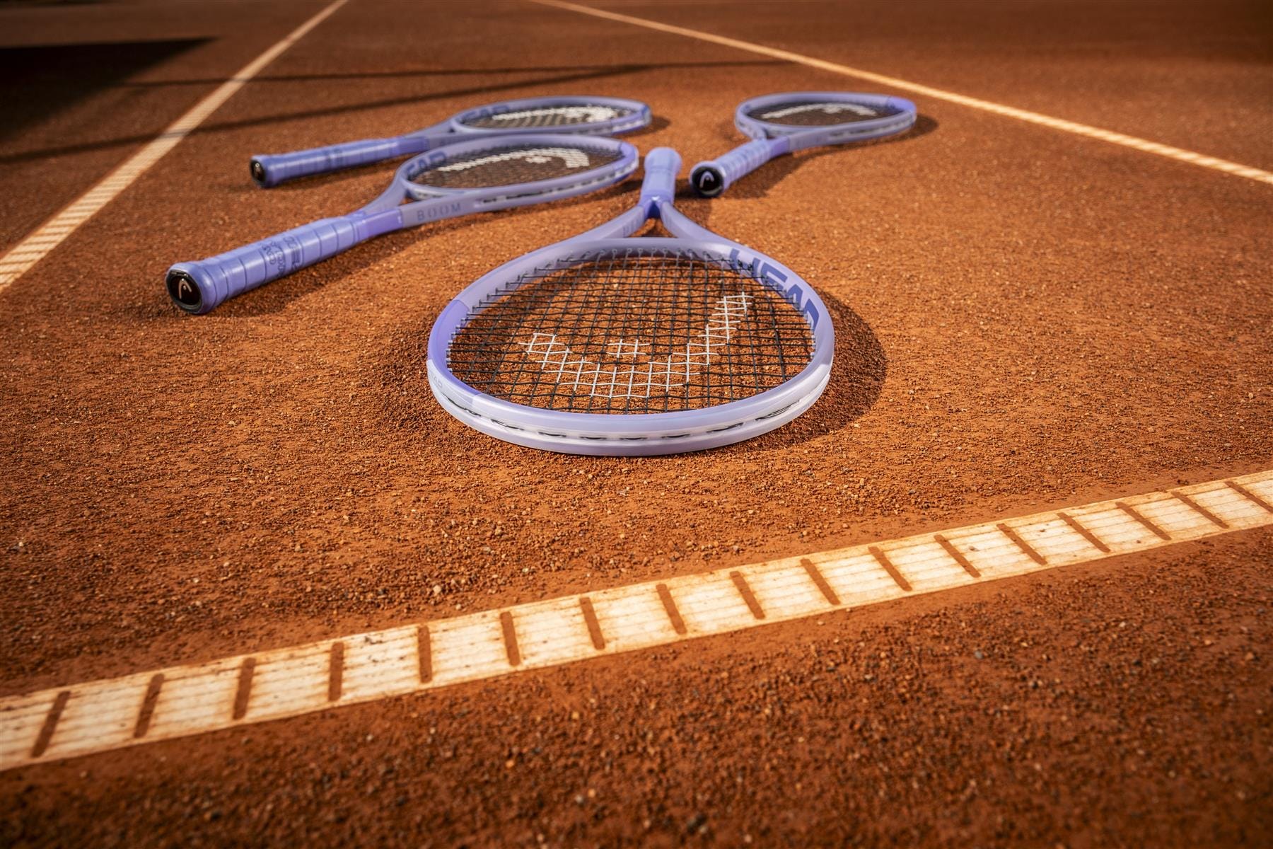 Four HEAD tennis rackets, including the HEAD Boom MP L Alternate 2026 Tennis Racket, lie near the baseline on the clay court. Distinct white lines highlight both power and touch on the reddish-brown surface.