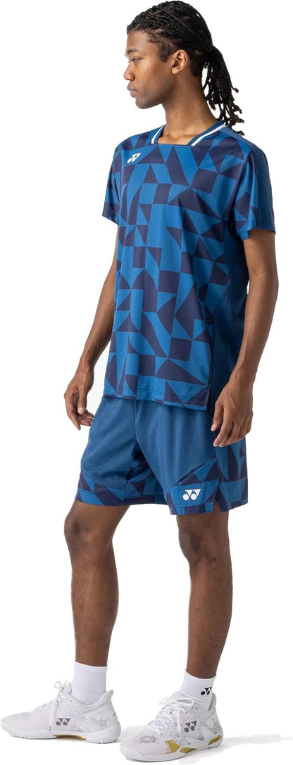 A person stands sideways wearing a blue geometric-patterned sports shirt and Yonex 15240YX Knit Mens Tennis Shorts in Indigo Blue, paired with white ankle socks and athletic shoes. They have braided hair and a neutral expression.