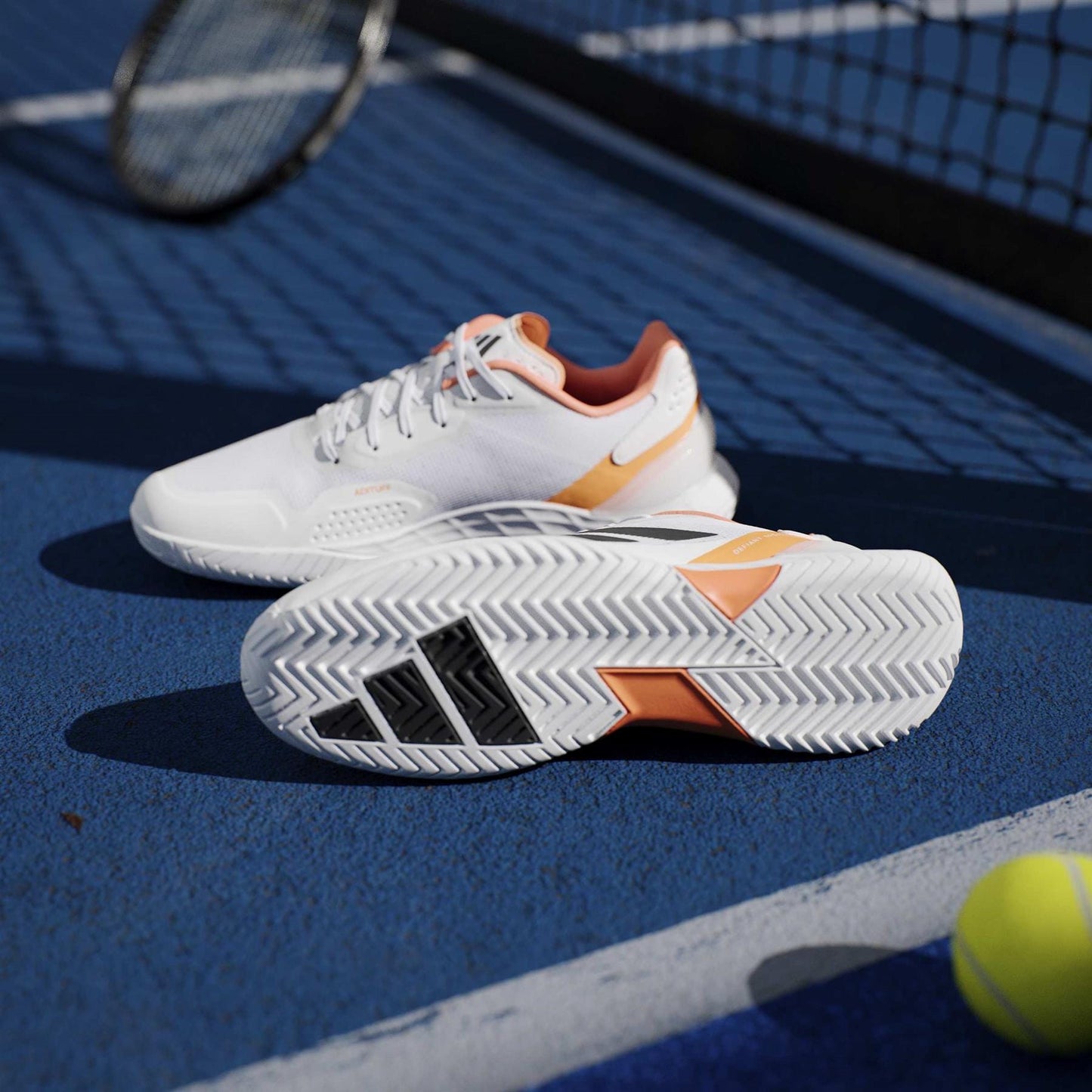 The adidas ADIDAS Defiant Speed 2 Men's Tennis Shoes in White/Orange are shown on a blue tennis court with a racket and yellow ball by the net, highlighting these lightweight men's shoes from adidas.