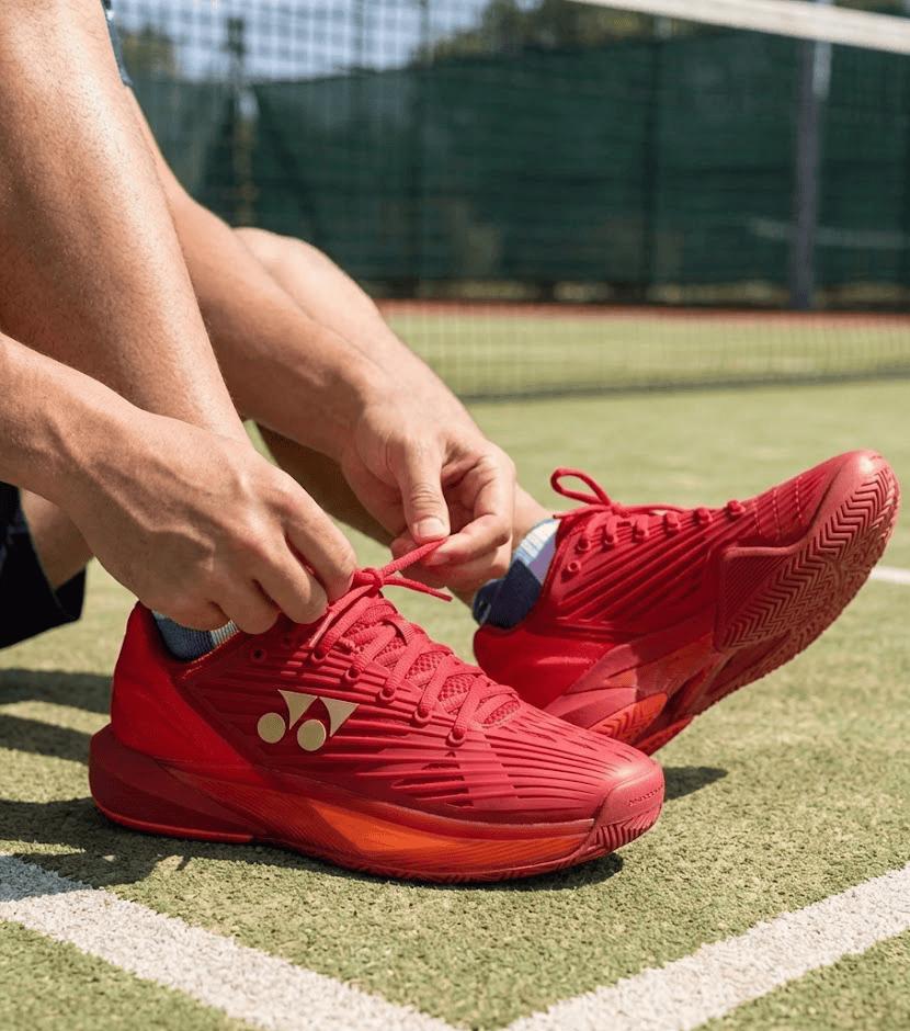 A person ties the laces of Yonex Power Cushion Eclipsion 5 Mens Tennis Shoes in Ruby Red, featuring Yonex’s Power Cushion technology, while sitting on an outdoor tennis court.