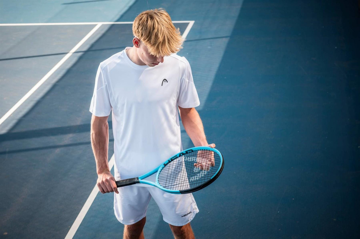 A tennis player in white stands on a blue court, looking down at his HEAD IG Boom XCEED Tennis Racket by HEAD as sunlight casts shadows across the court.