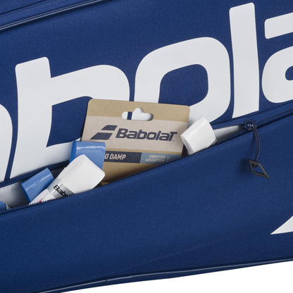 A close-up of the blue Babolat Court XS 3 Racket Tennis Bag's zippered pocket, partially open to reveal Babolat branded accessories, like a "Vibration Absorption" package and several white and blue items in this compact bag.