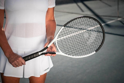 A tennis player in white holds the HEAD MX Attitude SUPRM Tennis Racket by HEAD on an outdoor court, with the net and shadows visible behind.
