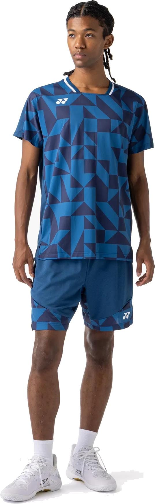 A young man with braided hair looks to the side, wearing a blue geometric athletic shirt and Yonex 15240YX Knit Mens Tennis Shorts in Indigo Blue, paired with white socks and sneakers, against a white background.