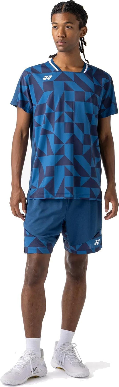 A young man with braided hair looks to the side, wearing a blue geometric athletic shirt and Yonex 15240YX Knit Mens Tennis Shorts in Indigo Blue, paired with white socks and sneakers, against a white background.