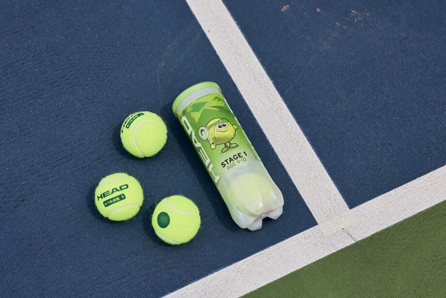 A can of HEAD 3B Stage 1 Tennis Balls - Green and three loose beginner tennis balls from HEAD rest on a blue and green tennis court near a white line.