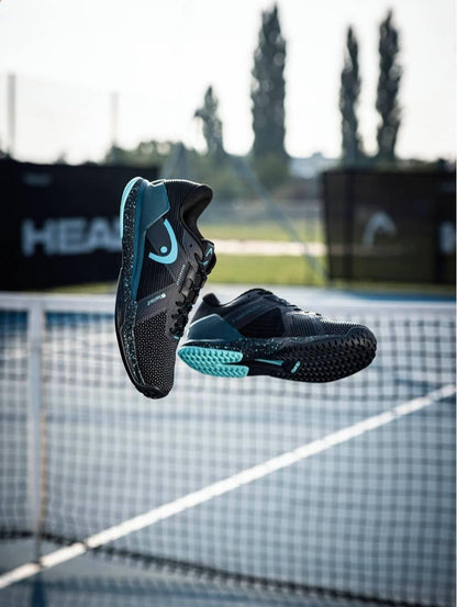 A pair of HEAD Sprint Pro 4.0 SF Men’s Tennis Shoes in black/teal are suspended above a tennis court, with blurred nets and trees in the background.