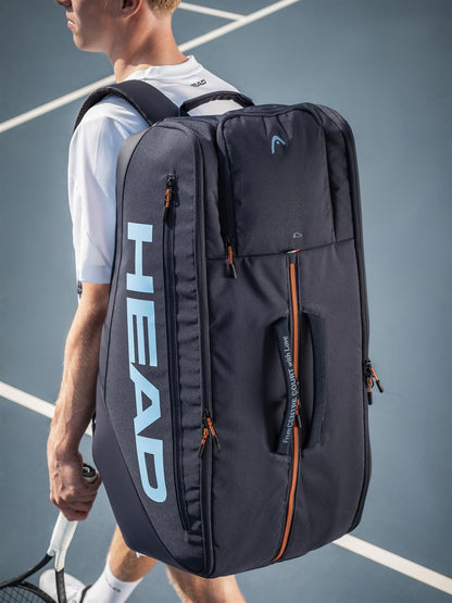 A tennis player stands on court, carrying the HEAD Pro 12 Racket Tennis Bag XL – Navy by HEAD, featuring blue lettering and orange zipper accents, designed to hold multiple rackets. The player also grips a tennis racket.