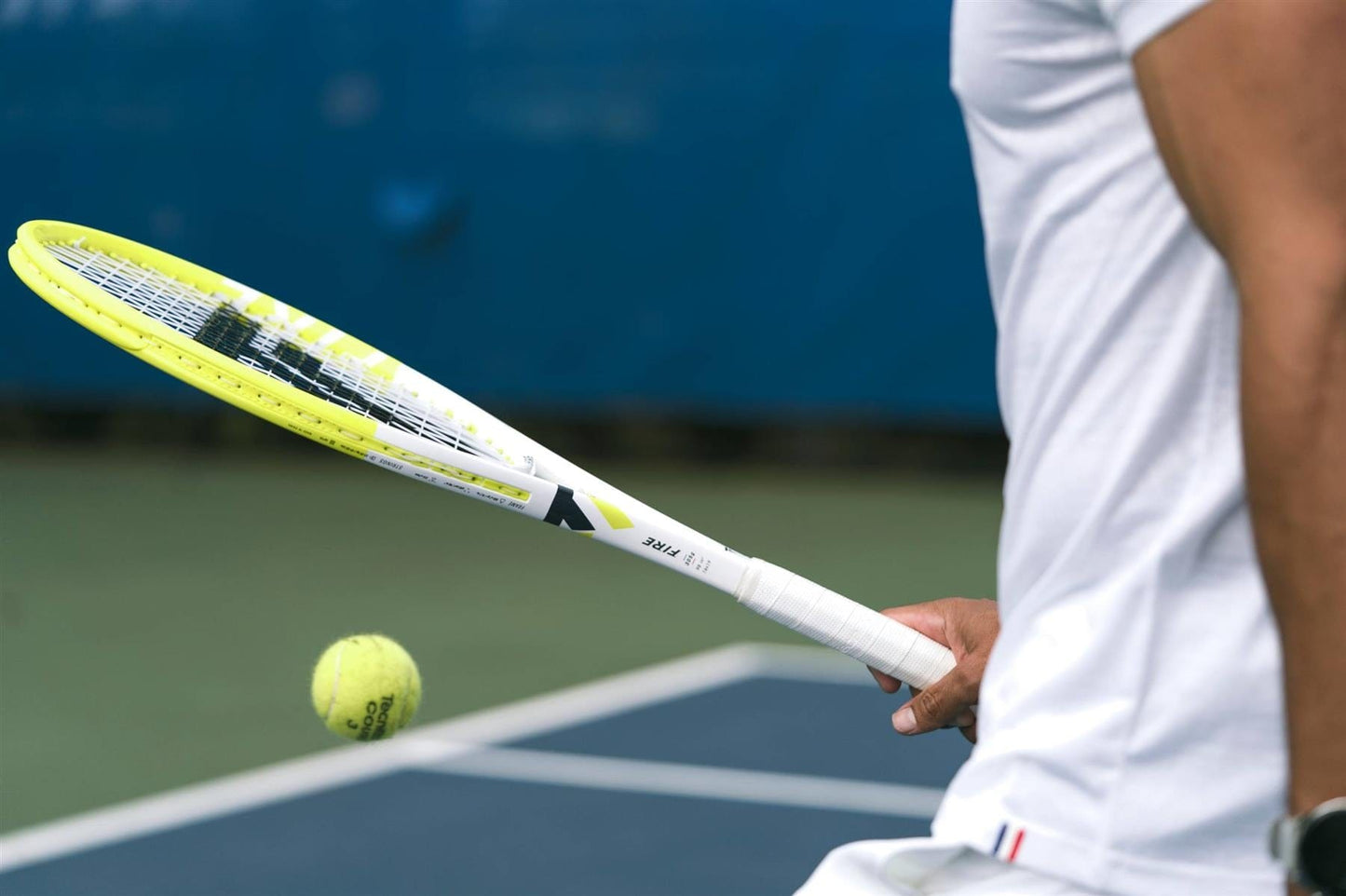 A person in white prepares to hit a yellow tennis ball with the Tecnifibre Fire 285 Tennis Racket by Tecnifibre, which features vibration dampening technology, on an outdoor court. The focus is on their arm, racket, and ball with a blue background.