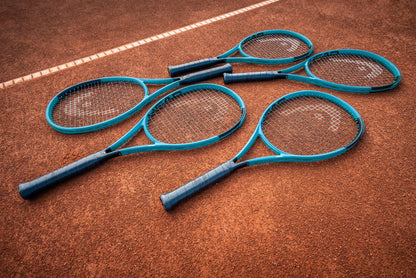 Six HEAD Boom MP L 2026 Tennis Rackets with blue frames and black handles are neatly arranged, overlapping on a clay court near a white line, ready to deliver effortless power.