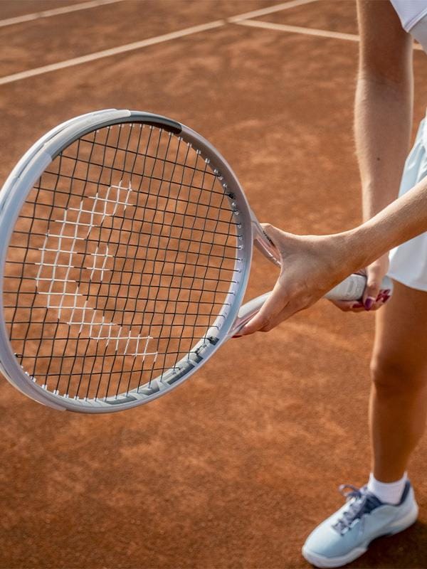 Dressed in white tennis gear, a player stands on a clay court holding the HEAD Squared Tennis Racket by HEAD. Only their lower body and one hand gripping the ultra-head light, dual-tube technology racket are visible.