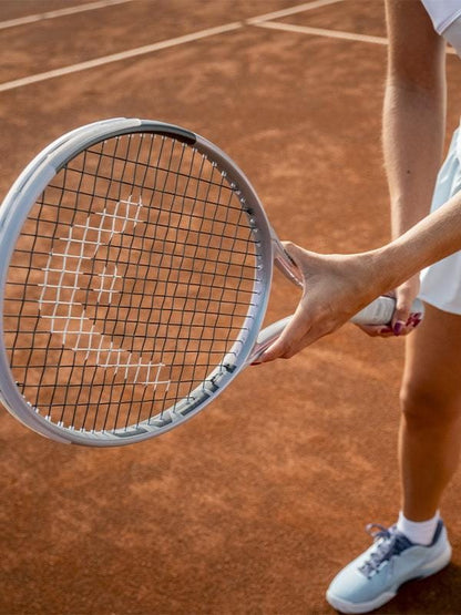 Dressed in white tennis gear, a player stands on a clay court holding the HEAD Squared Tennis Racket by HEAD. Only their lower body and one hand gripping the ultra-head light, dual-tube technology racket are visible.