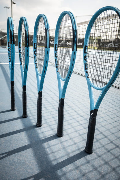 Five HEAD Boom MP L 2026 Tennis Rackets stand upright on a blue tennis court near the net, casting shadows in the sunlight and promising effortless power with every swing.