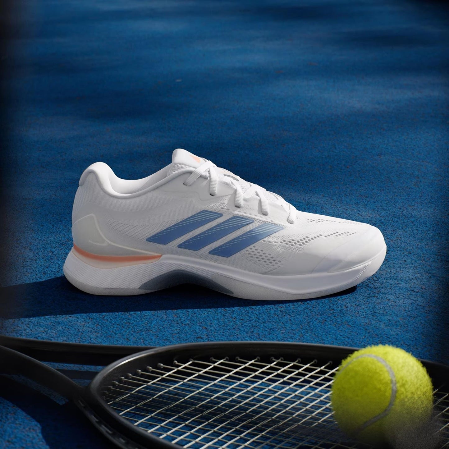 A pair of adidas ADIDAS Avacourt 3 Women’s Tennis Shoes in Core White, Halo Blue, and Beam Orange sits on a blue tennis court beside a black racket and a yellow ball.