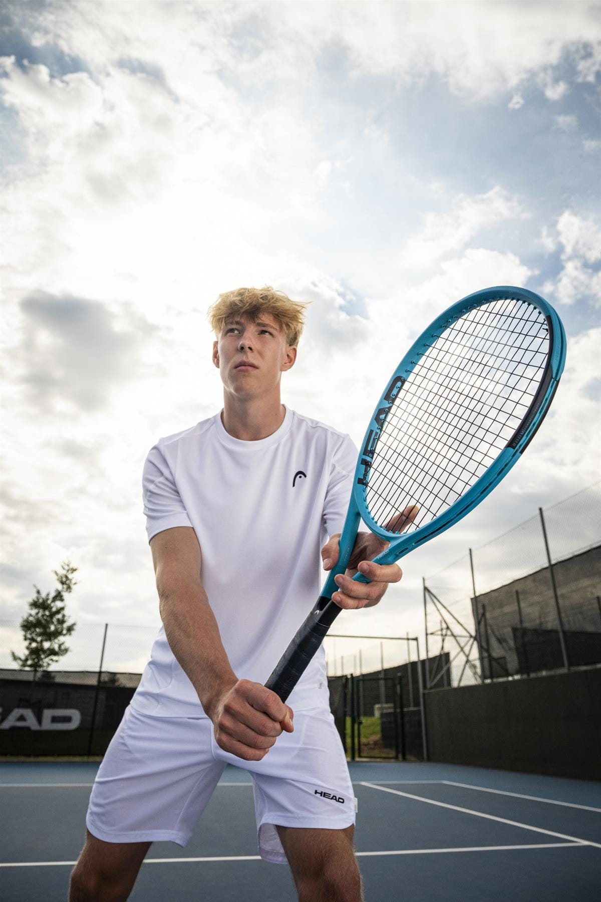 A young man in a white shirt and shorts readies to strike a tennis ball with the HEAD Boom MP 2026 Tennis Racket on an outdoor court under cloudy skies, showcasing the power and precision of HEAD’s latest racquet.