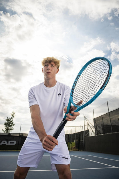 A young man in a white shirt and shorts readies to strike a tennis ball with the HEAD Boom MP 2026 Tennis Racket on an outdoor court under cloudy skies, showcasing the power and precision of HEAD’s latest racquet.