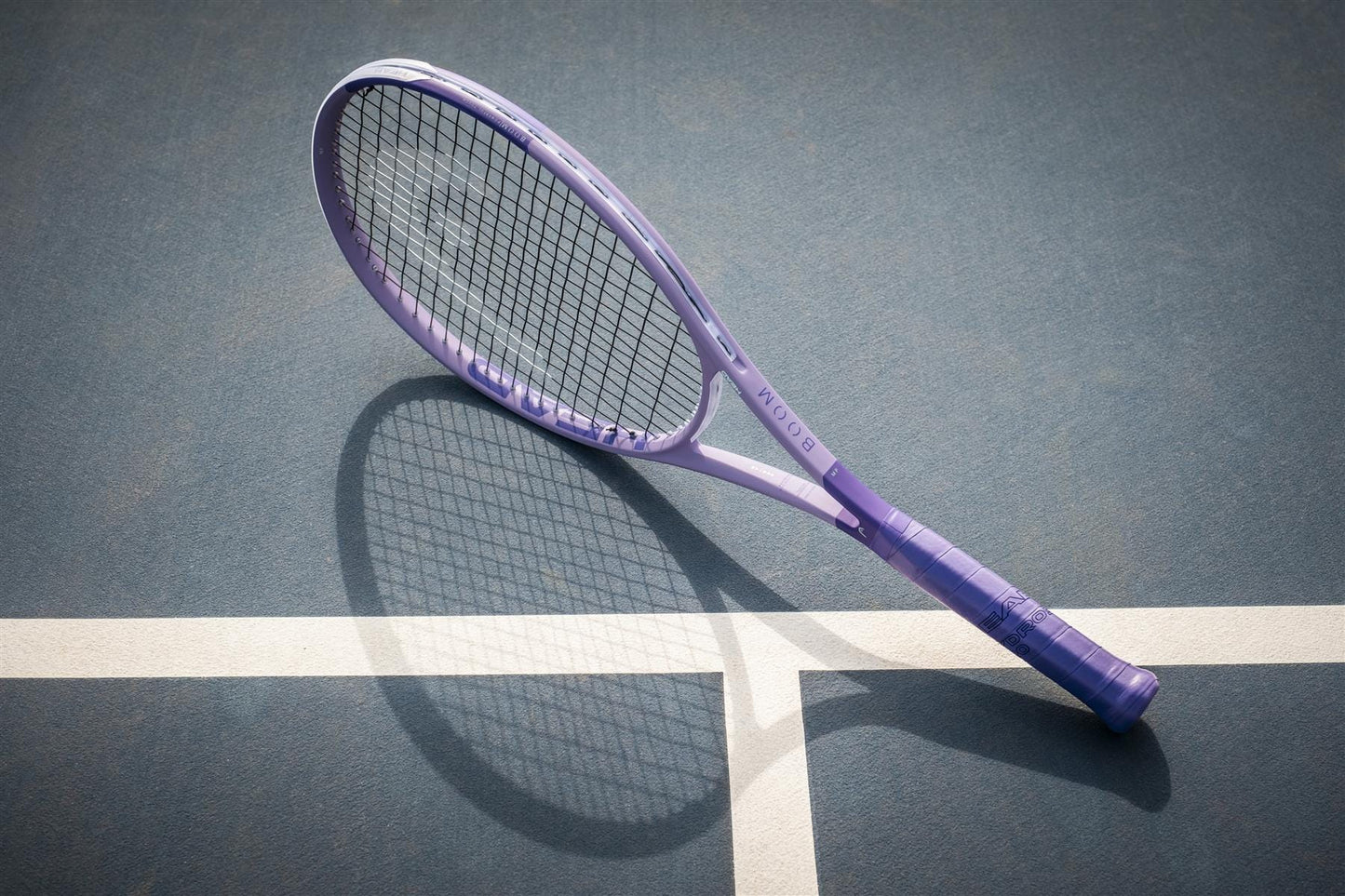 A HEAD Boom MP Alternate 2026 Tennis Racket in purple rests on a blue court beside a white line, its shadow emphasizing the dynamic power and performance that HEAD delivers.