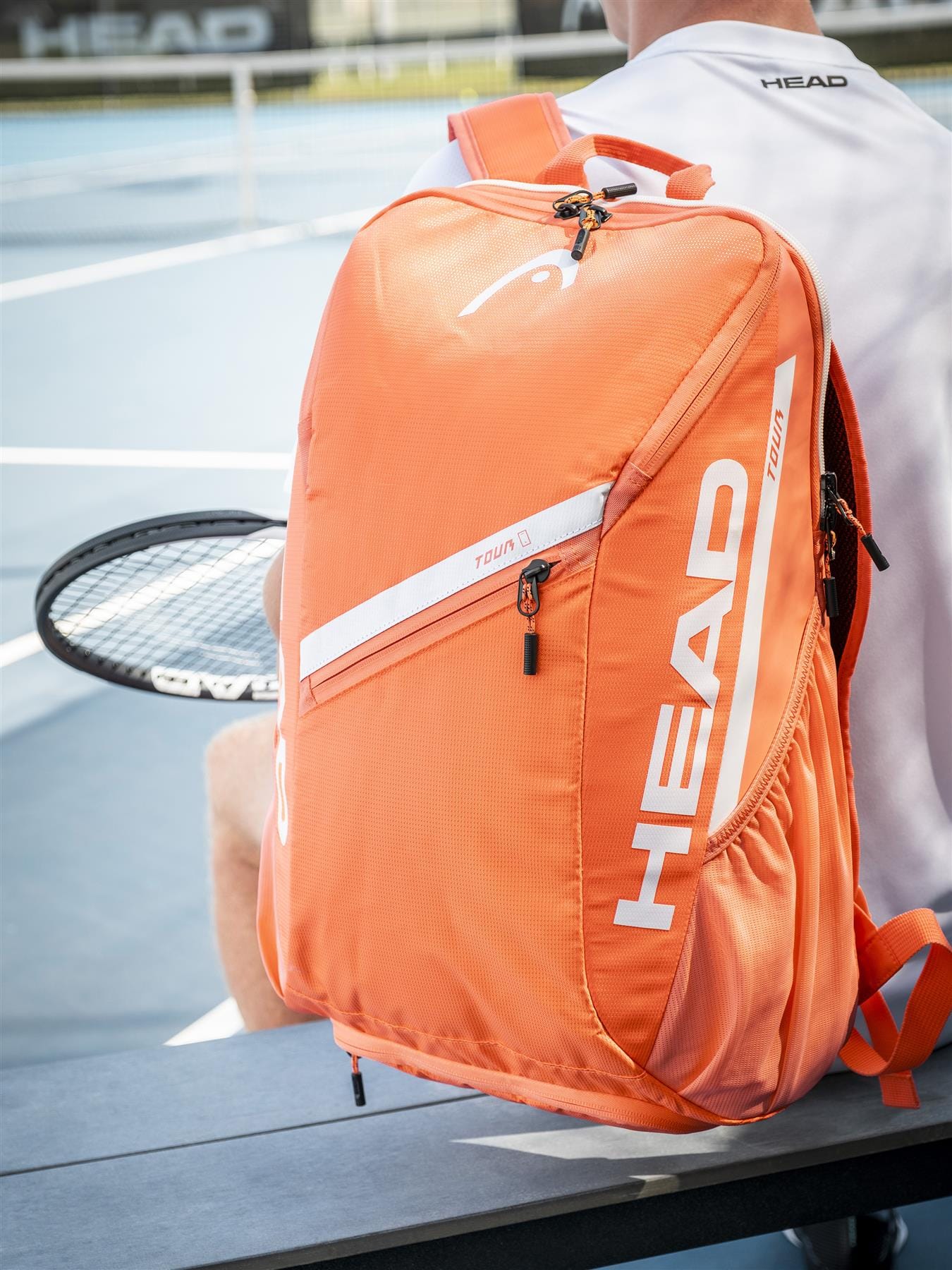 An orange HEAD Tour 25L Tennis Backpack rests on a bench beside a player holding a racket on a blue outdoor tennis court, with the HEAD logo and "Tour Team" text visible on the stylish bag.