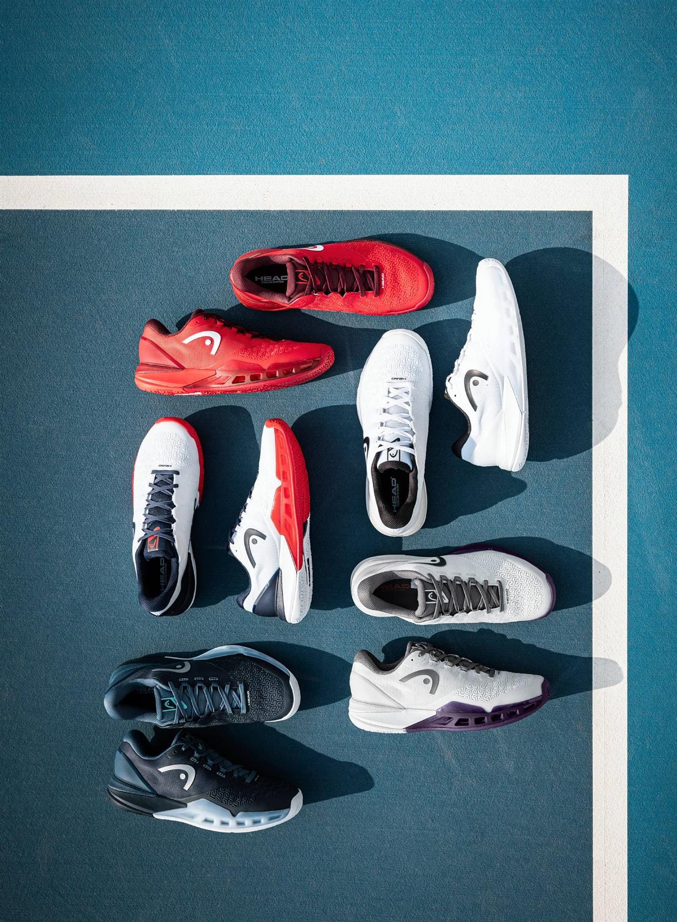 Eight HEAD Revolt Pro 5.0 Men’s Tennis Shoes in white and navy, from HEAD, are neatly arranged on a blue tennis court by a white line, highlighting stylish options for baseline players.