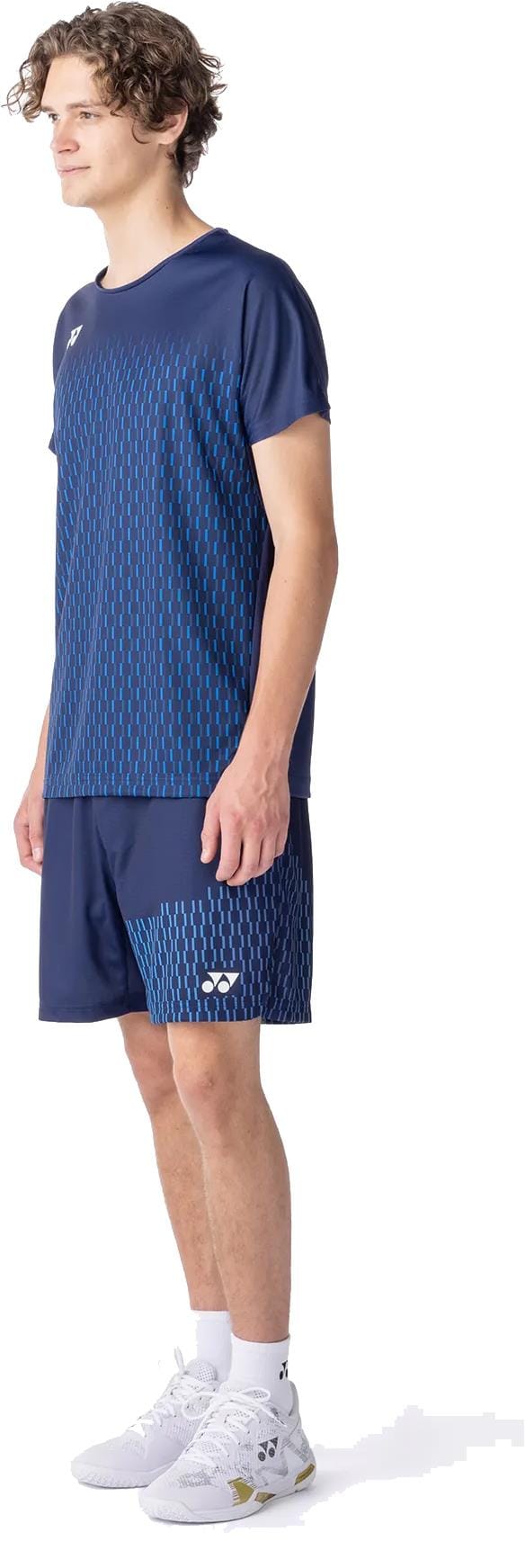 A young man wears the Yonex 10713EX Crew Neck Men's Tennis T-Shirt in Dark Navy, paired with blue dotted shorts, white crew socks, and white sports shoes. He stands sideways, looking slightly forward.