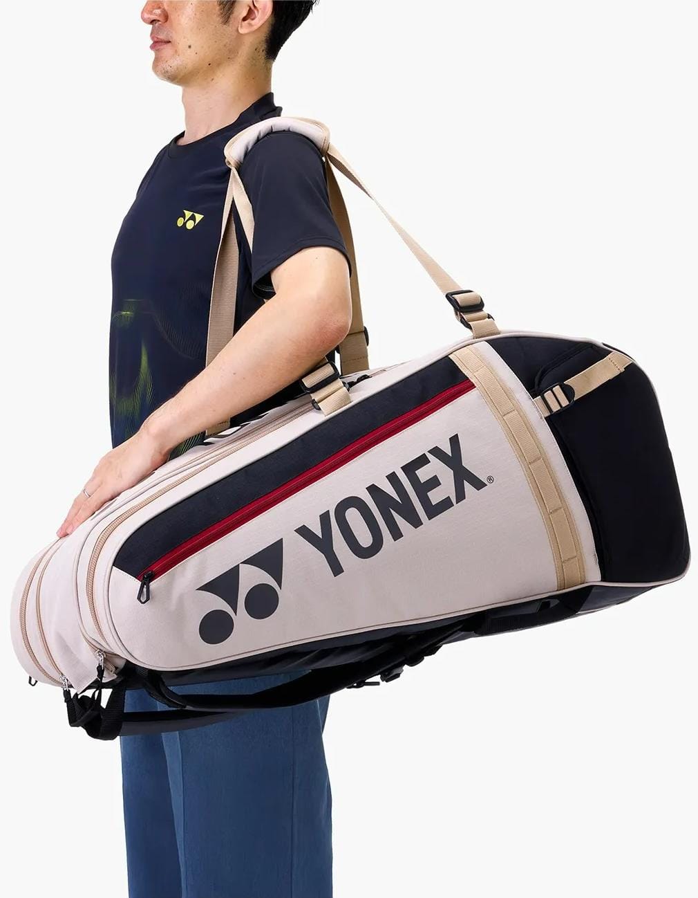 A person in a dark Yonex shirt carries the Yonex BA72629EX Gearlogic 9 Racket Tennis Bag in beige, a spacious Yonex bag with a bold logo that easily fits rackets and gear.