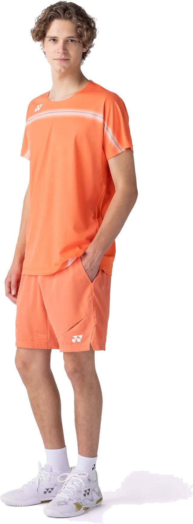 A young man with curly hair models the Yonex 10711YX Crew Neck Mens Tennis T-Shirt in Cyber Orange, paired with matching shorts, white socks, and sports shoes. He stands confidently against a white background, showcasing sleek Yonex sportswear.