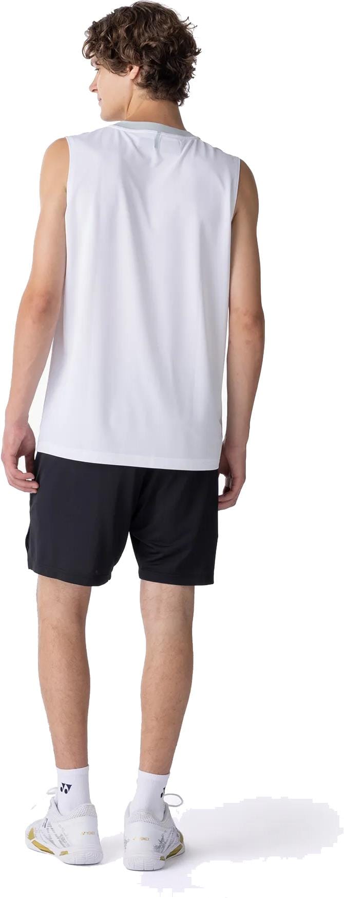 A young man, facing away, wears a sleeveless white shirt and Yonex 15239YX Knit Mens Tennis Shorts in black. He pairs them with white crew socks and athletic shoes. His curly hair and relaxed posture complete the look.