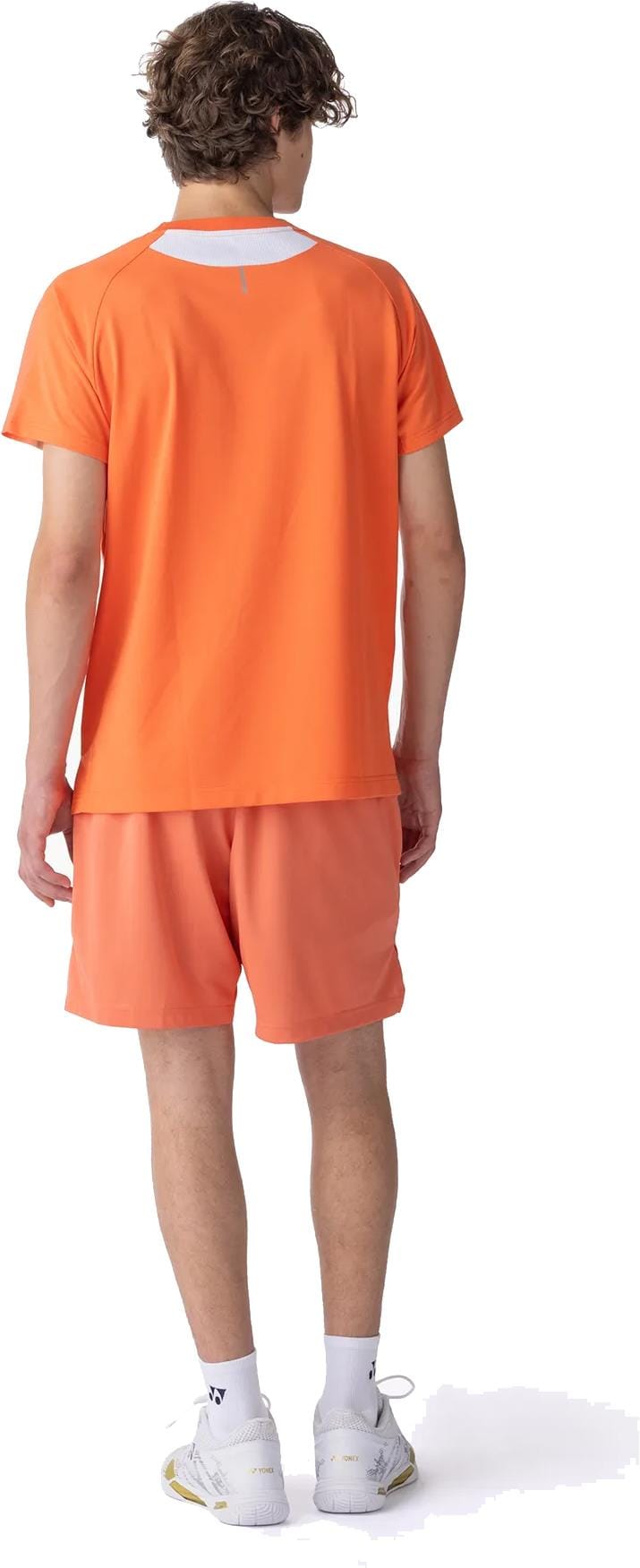 A young man faces away, wearing a Yonex 10711YX Crew Neck Men's Tennis T-Shirt in Cyber Orange and matching shorts, with white socks and athletic shoes, set against a white background.
