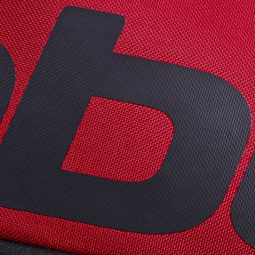 Close-up of a dark grey letter "b" printed on a textured, bright red fabric background, reminiscent of the bold branding found on the Babolat Court L 9 Racket Tennis Bag in Burgundy and Black.