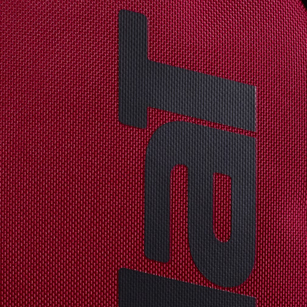 A close-up of the black Babolat logo, featuring the letter "T" and a stylized "R," printed on the textured burgundy fabric of the Babolat Court Hero Tennis Backpack—crafted from recycled polyester for a premium look and feel.