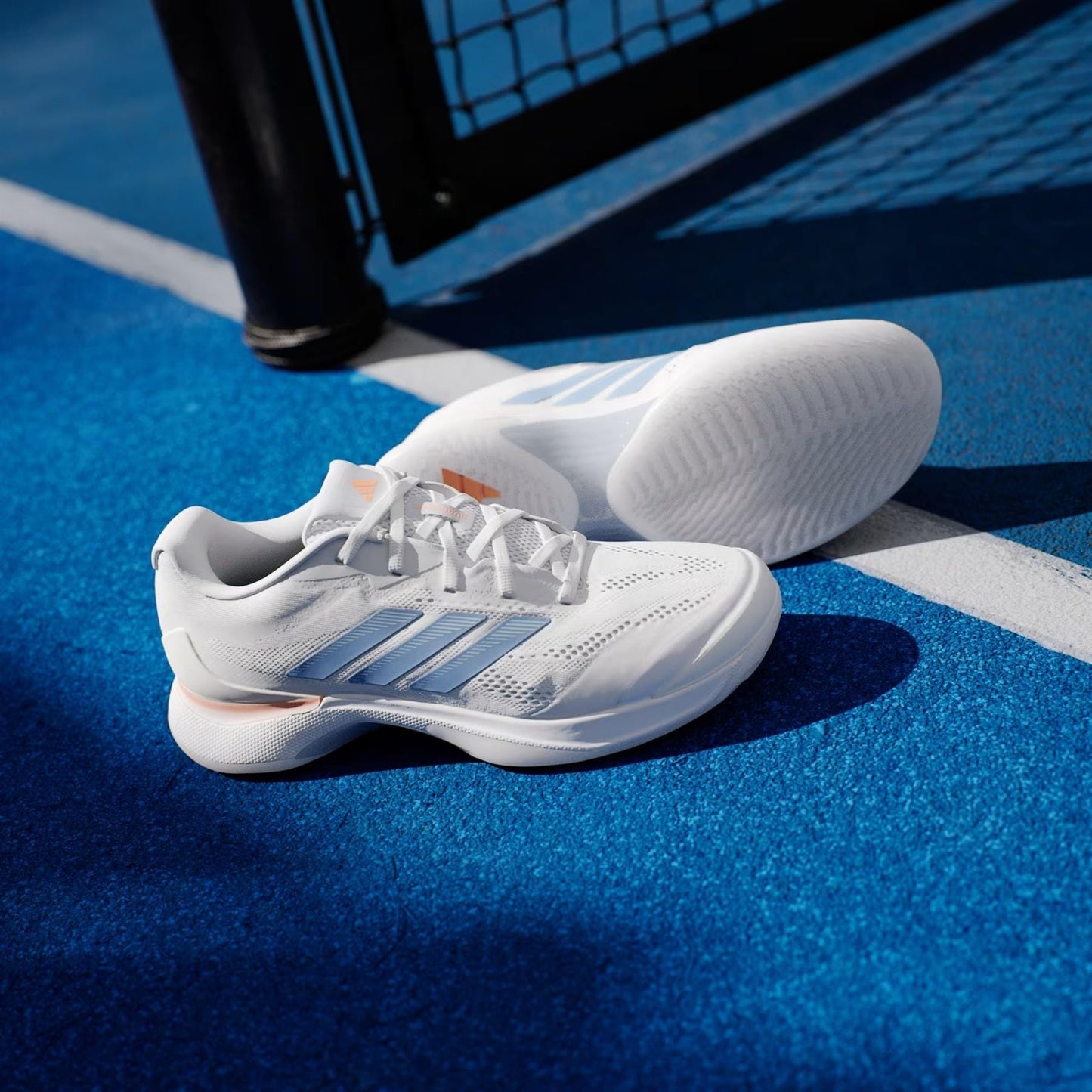 A pair of adidas Avacourt 3 Women's Tennis Shoes in Core White, Halo Blue, and Beam Orange are displayed on a bright blue tennis court beside a black net post.