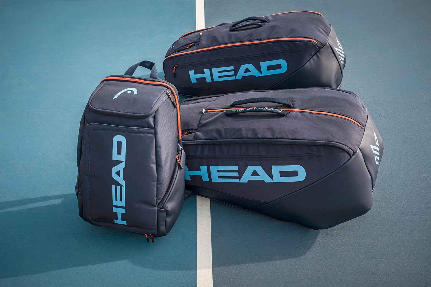 Three HEAD Pro 9 Racket Tennis Bag L - Navy by HEAD, featuring blue logos and orange accents, are placed on a blue tennis court near a white line.