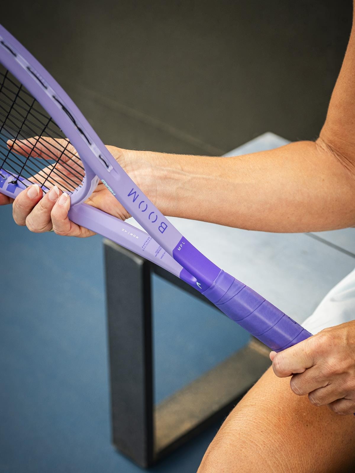 A person sits on a bench gripping the handle of a purple HEAD Boom MP L Alternate 2026 Tennis Racket, one hand on the racket and the other adjusting the grip for optimal power and touch.