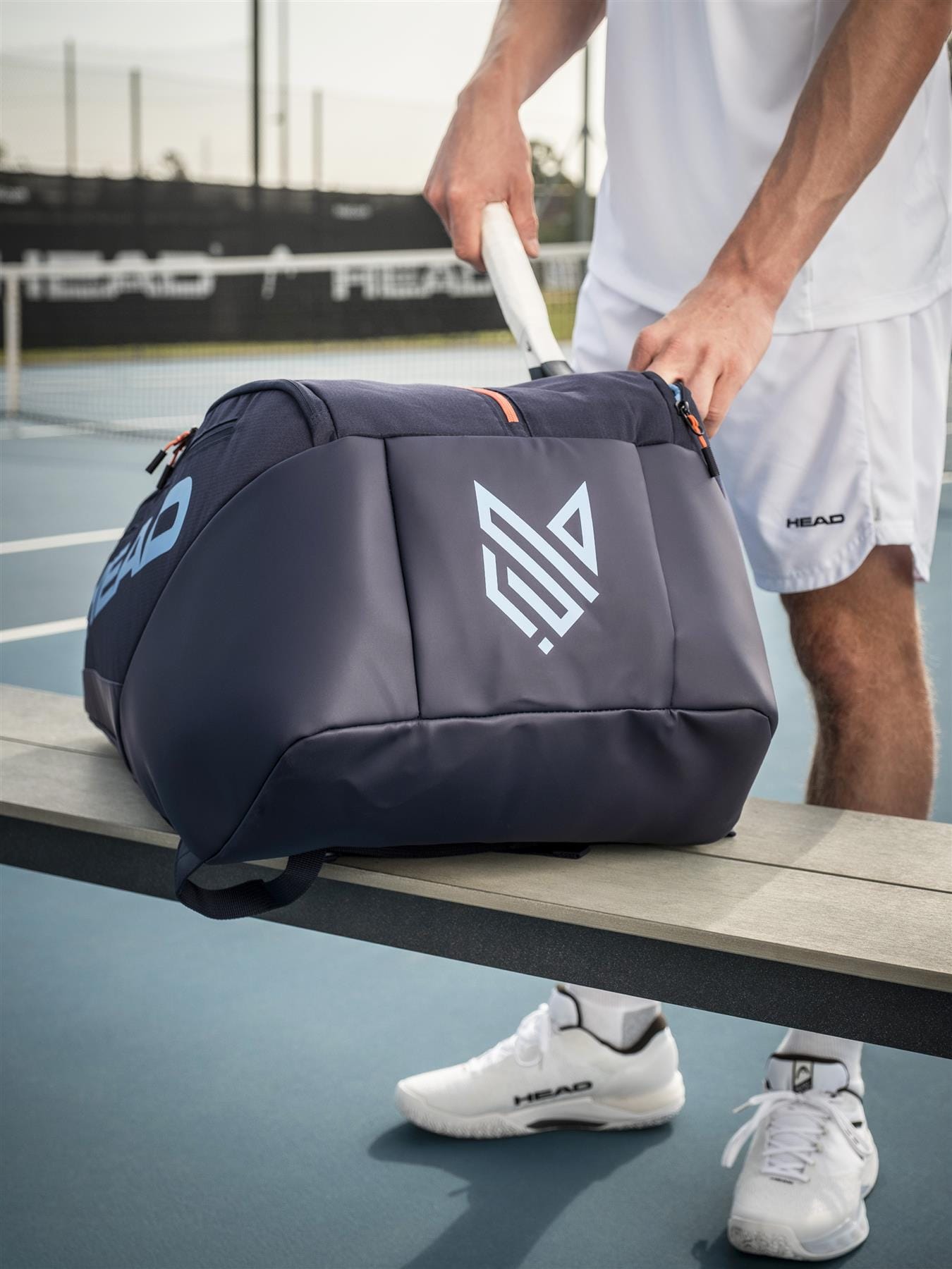 A tennis player in white sportswear stands by an outdoor court bench, reaching into a HEAD Pro 12 Racket Tennis Bag XL - Navy by HEAD, with rackets and the court visible in the background.