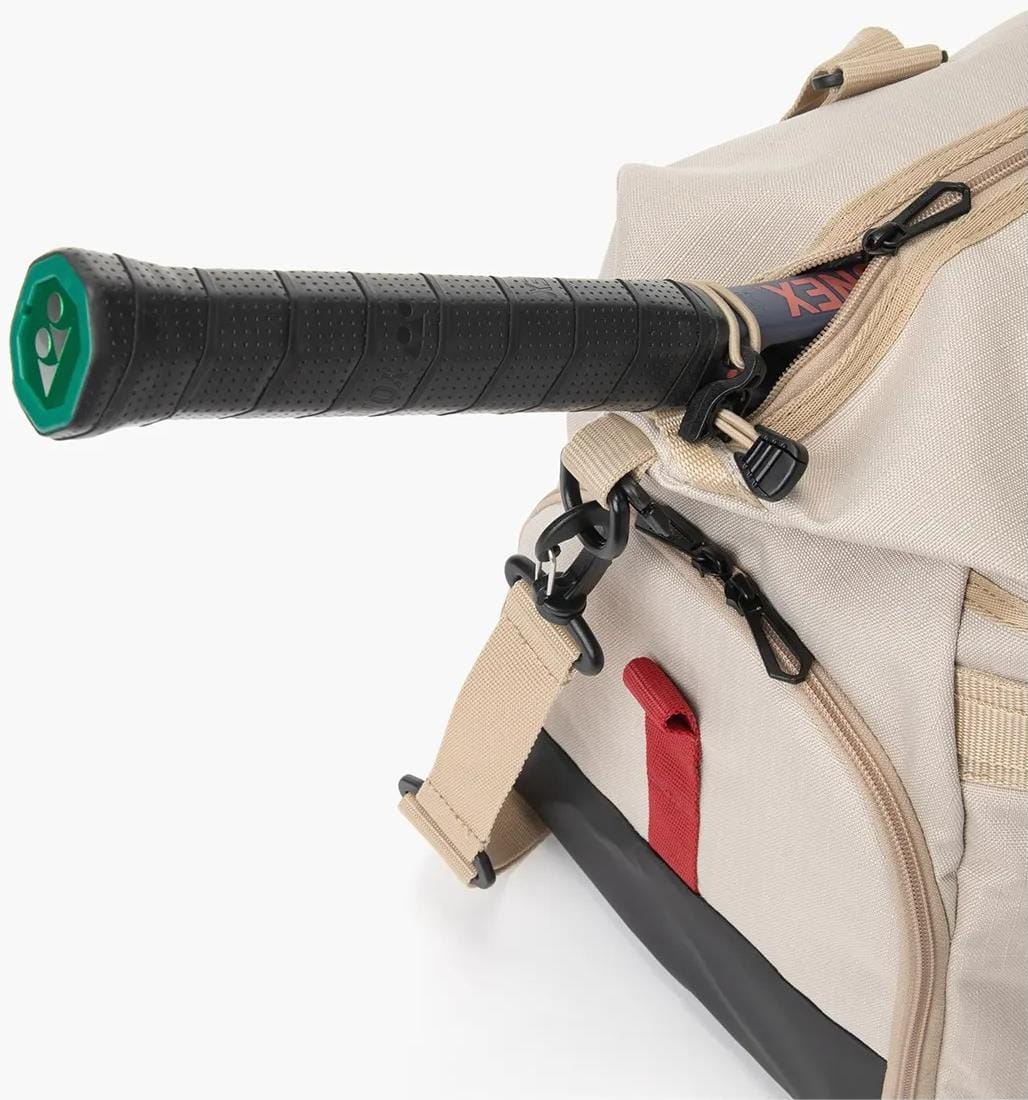 A close-up of the Yonex BA72611EX Gearlogic Tennis Boston Bag in beige shows a black and green racket handle in the side pocket. The recycled polyester bag features black clips with red and tan accents.