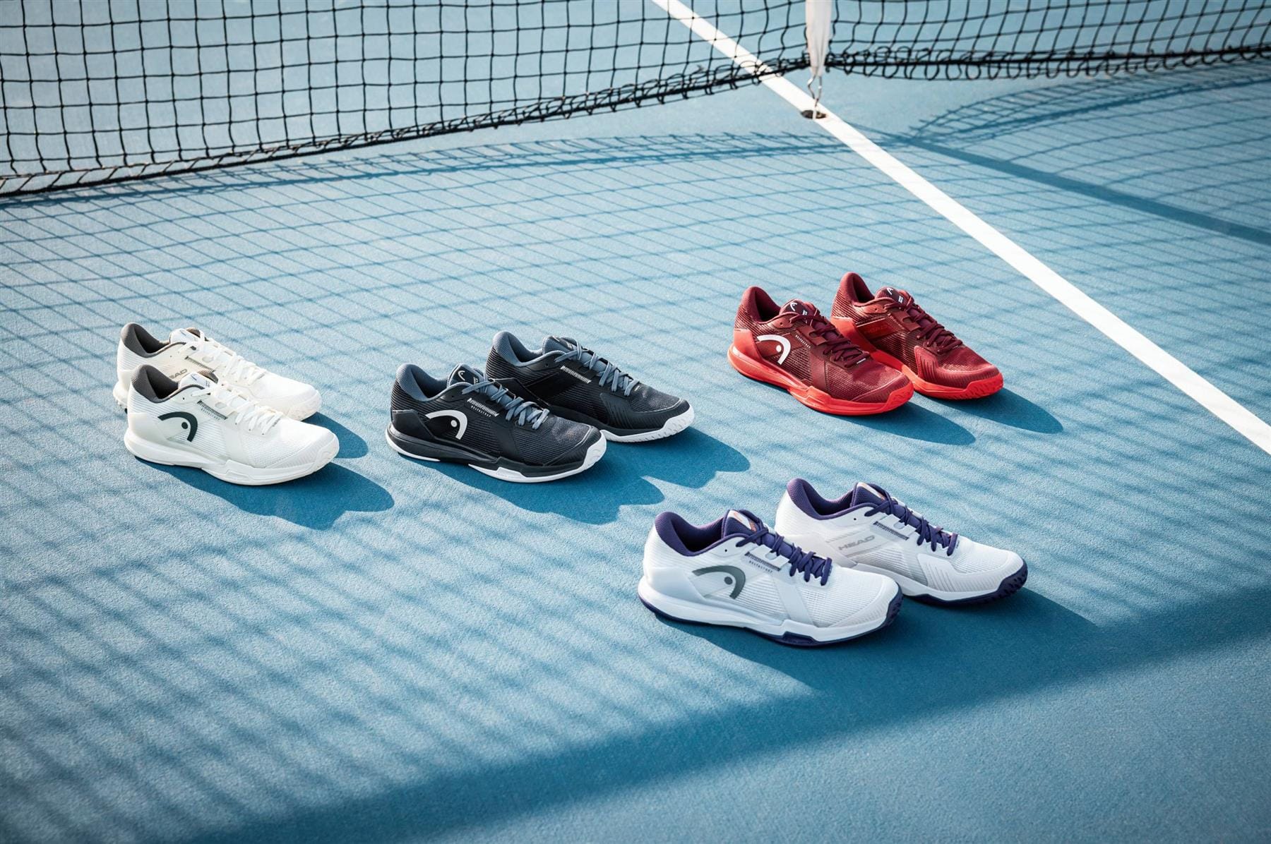 Four pairs of HEAD Sprint Pro 4.0 Mens Tennis Shoes in Chalk White/Dark Grey are lined up on a blue tennis court near the net, showcasing their brand's signature breathable and lightweight design.